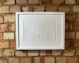 May include: A white framed print of a white paper cut-out of a building. The building is a large, multi-story structure with a central archway and multiple windows. The print is on a brick wall background.