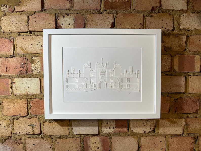 May include: A white framed print of a white paper cut-out of a building. The building is a large, multi-story structure with a central archway and multiple windows. The print is on a brick wall background.