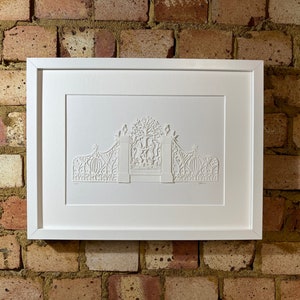 May include: A white framed print of a white paper cut design of a gate with a tree and intricate details.