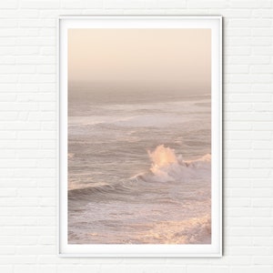 May include: A photograph of a wave crashing in the ocean at sunset. The water is a light blue and the sky is a soft pink.