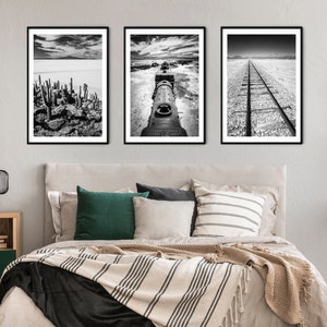 May include: Three framed black and white landscape prints. The images depict a cactus landscape, a train engine, and a railroad track. The artwork is displayed above a bed with pillows and a striped blanket.