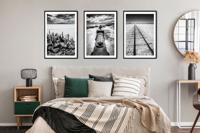 May include: Three framed black and white landscape prints. The images depict a cactus landscape, a train engine, and a railroad track. The artwork is displayed above a bed with pillows and a striped blanket.