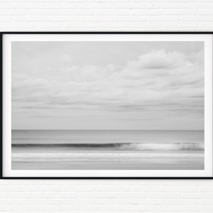 May include: A black and white photograph of a wave crashing on a beach. The sky is cloudy and the water is calm.