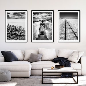 May include: Three framed black and white landscape photographs. The images depict a desert scene with cacti, a train engine, and a railroad track. The artwork is displayed above a white sectional sofa in a living room setting.