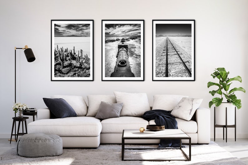 May include: Three framed black and white landscape photographs. The images depict a desert scene with cacti, a train engine, and a railroad track. The artwork is displayed above a white sectional sofa in a living room setting.