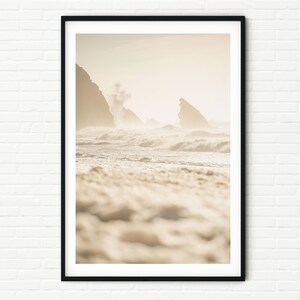 May include: A framed print depicting a coastal landscape. The photograph shows a beach with waves breaking on the shore. The sky is misty, and the colour scheme is soft, with beige and off-white hues. The print is presented in a black frame.