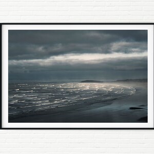 Puede incluir: Una fotografía de una playa con olas rompiendo en la orilla. El cielo está cubierto de nubes oscuras, pero hay un rayo de luz que se abre paso a través de las nubes.