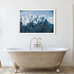 May include: A white freestanding bathtub with a gold faucet and shower head. The bathtub is in a bathroom with a white tiled floor and a white wall. A framed photograph of a glacier is hanging on the wall above the bathtub.