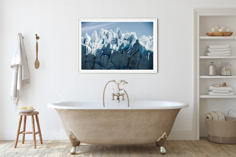 May include: A white freestanding bathtub with a gold faucet and shower head. The bathtub is in a bathroom with a white tiled floor and a white wall. A framed photograph of a glacier is hanging on the wall above the bathtub.