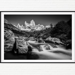 May include: Black and white framed landscape photograph of a mountain range with snow-capped peaks. A river flows through the foreground, with blurred water creating a sense of movement. The image is set against a clear sky.