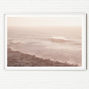 May include: A photograph of ocean waves crashing on a sandy beach. The water is a soft pink color, and the sky is a light blue. The waves are breaking in the distance, and the sun is shining brightly.