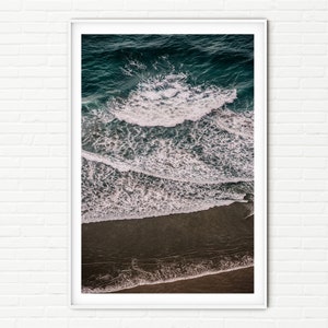 Puede incluir: Una vista aérea de olas del océano rompiendo en una playa de arena. El agua es de un azul profundo y las olas son blancas con espuma. La arena es de un marrón claro.