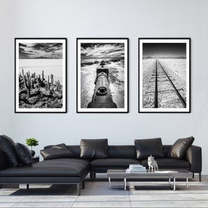 May include: Three framed black and white landscape photographs. The images depict a desert scene with cacti, a train, and a railway track. The artwork is displayed above a black leather sectional sofa in a modern living room.