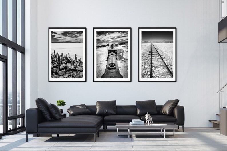 May include: Three framed black and white landscape photographs. The images depict a desert scene with cacti, a train, and a railway track. The artwork is displayed above a black leather sectional sofa in a modern living room.