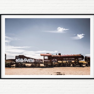Könnte beinhalten: Ein rostiger, mit Graffiti besprühtter Eisenbahnwagen steht verlassen in einer Wüstenlandschaft. Der Himmel ist blau mit weißen Wolken. Der Wagen ist mit Graffiti bedeckt, darunter die Worte "Asi es la vida", was übersetzt "So ist das Leben" bedeutet.