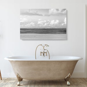 May include: A black and white photograph of a beach scene with a cloudy sky and a calm ocean. The photo is hanging on a white wall above a freestanding bathtub.