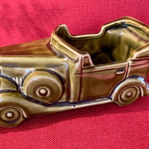 May include: A ceramic planter shaped like a vintage car, in shades of olive green and brown. The car planter features detailed wheels, a convertible top, and a classic design, perfect for small plants or decorative use.