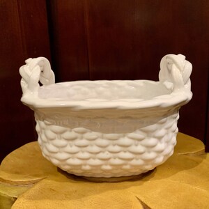 May include: A white ceramic basket with a textured, woven-like pattern. The basket has two braided handles and a rounded shape. The interior is smooth, and the overall design is clean and simple, suitable for various decorative uses.