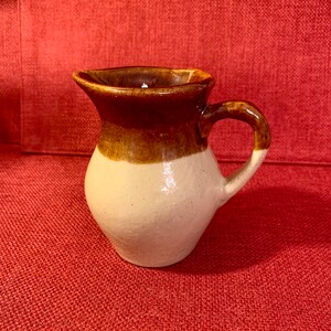 May include: A small, handcrafted ceramic pitcher with a rounded body and a handle. The top and handle are a rich brown, contrasting with the off-white lower half. The pitcher has a spout for easy pouring and is likely used for serving beverages.