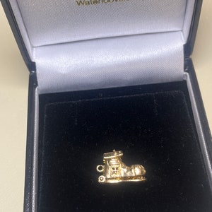 May include: A gold boot charm, presented in a black velvet-lined jewellery box. The charm features detailed design elements, including a window and decorative features. The box is black with a white satin lining. The shop name is visible.