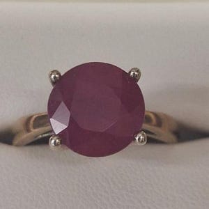 May include: A gold ring with a large, round, faceted ruby gemstone. The ruby is a deep red colour. The ring has a classic four-prong setting.