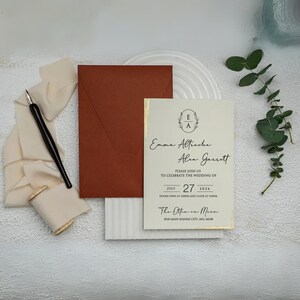 May include: Wedding invitation with a cream-colored card featuring gold edging and the names Emma Altreche and Alex Garrett. A rust-colored envelope, a black pen, and cream-colored ribbon are arranged on a textured white surface. The invitation includes the date July 27, 2026.