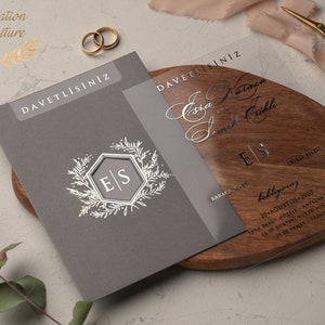 May include: A gray invitation card with the text "DAVETLİSİNİZ" in gold lettering. The card features a silver monogram with the initials "E|S" surrounded by a floral design. The card is on a wooden surface with a clear acrylic overlay with the text "DAVETLİSİNİZ" in silver lettering. The overlay also features the names "Eda Karaca" and "Samet &Ccedil;aklı" in cursive script, along with the date "20 Ağustos 2023" and the location "Ramada Resort Antalya Sara" in black lettering.
