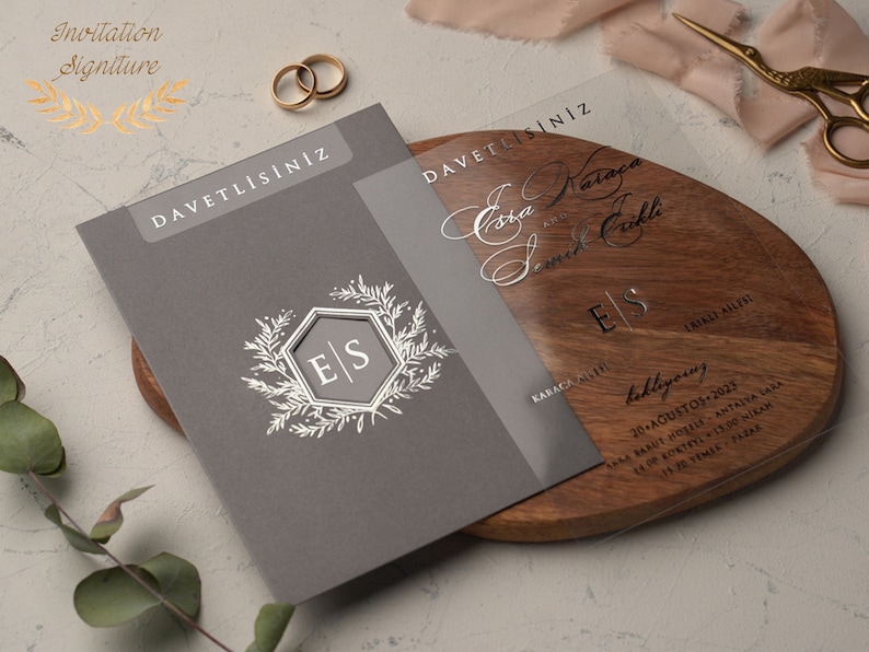 May include: A gray invitation card with the text "DAVETLİSİNİZ" in gold lettering. The card features a silver monogram with the initials "E|S" surrounded by a floral design. The card is on a wooden surface with a clear acrylic overlay with the text "DAVETLİSİNİZ" in silver lettering. The overlay also features the names "Eda Karaca" and "Samet &Ccedil;aklı" in cursive script, along with the date "20 Ağustos 2023" and the location "Ramada Resort Antalya Sara" in black lettering.