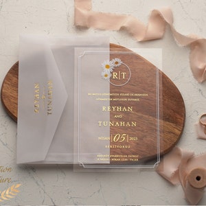 Könnte beinhalten: Eine transparente Acryl-Hochzeitseinladung mit goldenen Lettern und einem floralen Design. Die Einladung lautet "REYHAN UND TUNAHAN" und enthält das Datum "NISAN 05 2023".