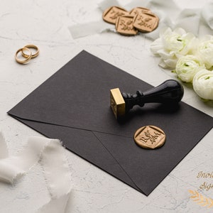May include: A black envelope with a gold wax seal and a black wax stamp with a wooden handle. The wax seal has the initials "R & M" in a decorative font. The envelope is on a white surface with white ribbon and white flowers.