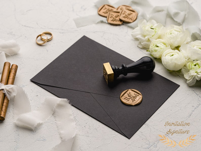 May include: A black envelope with a gold wax seal and a black wax stamp with a wooden handle. The wax seal has the initials "R & M" in a decorative font. The envelope is on a white surface with white ribbon and white flowers.