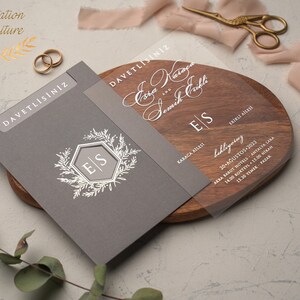 May include: A gray and clear wedding invitation with the names "Eda" and "Semih" written in cursive. The invitation includes the date, time, and location of the wedding ceremony and reception. The invitation also features a floral design with the initials "E" and "S" in a hexagon.