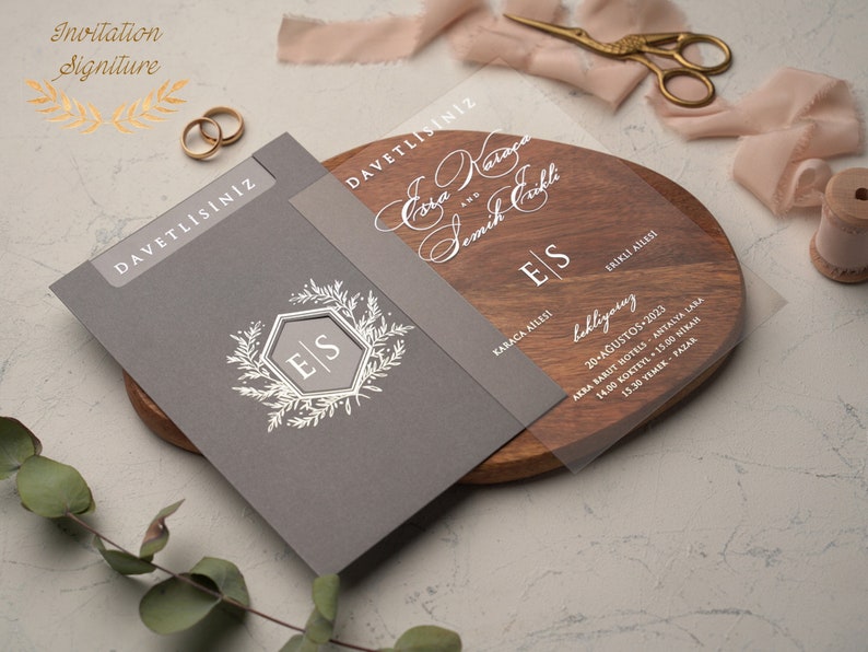 May include: A gray and clear wedding invitation with the names "Eda" and "Semih" written in cursive. The invitation includes the date, time, and location of the wedding ceremony and reception. The invitation also features a floral design with the initials "E" and "S" in a hexagon.