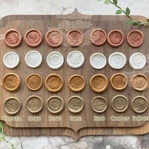 May include: A wooden board with 4 rows of 8 wax seals, each with a different design. The wax seals are in 4 colors: rose gold, pearl, caramel, and gold. The designs include a clover, spike, ring, rose, bird, crown, and flower.