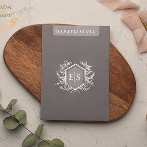 May include: A gray invitation card with the text "DAVETLISINIZ" printed in white at the top. The card features a silver hexagonal monogram with the initials "E|S" surrounded by a wreath of silver leaves.