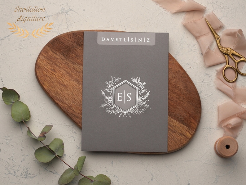 May include: A gray invitation card with the text "DAVETLISINIZ" printed in white at the top. The card features a silver hexagonal monogram with the initials "E|S" surrounded by a wreath of silver leaves.