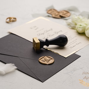 May include: A black envelope with a gold wax seal and a black and gold stamp. The wax seal has the initials "R & M" in gold lettering. The stamp is a traditional wooden stamp with a black handle.