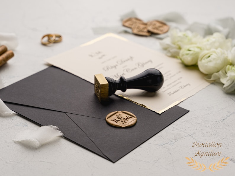 May include: A black envelope with a gold wax seal and a black and gold stamp. The wax seal has the initials "R & M" in gold lettering. The stamp is a traditional wooden stamp with a black handle.