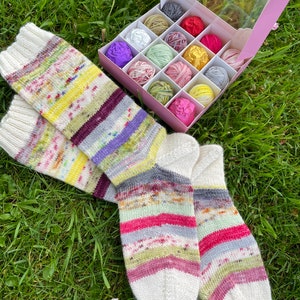May include: A pair of colourful striped knitted socks with white toes and heels. The socks are lying on a bed of green grass with a box of colourful yarn balls in the background.