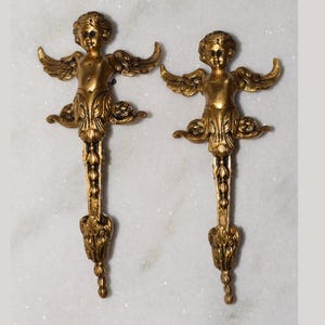 Peut inclure: Deux appliques murales antiques en forme de chérubin dorés. Chaque chérubin a des ailes déployées et est fixé à une base décorative et allongée. Les appliques sont en métal et sont présentées sur un fond de marbre blanc.