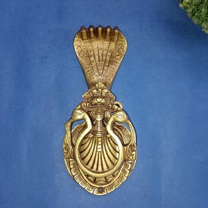 May include: A brass door knocker featuring a cobra head design at the top and a decorative oval base with two swan-like figures. The knocker is a warm gold color against a blue background.