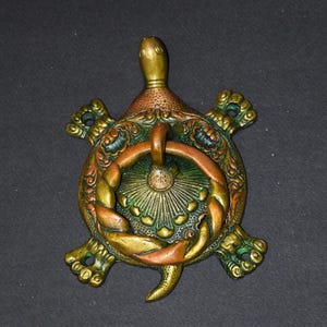 May include: A decorative turtle-shaped door knocker. The knocker is made of metal with a mix of green, gold, and copper tones. The turtle has a ring for knocking and intricate details on its shell and flippers.