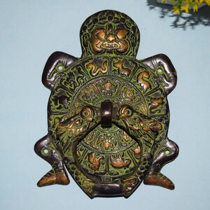 May include: Antique bronze door knocker with a detailed design. The knocker features a central circular element with intricate patterns and a handle, flanked by two dragon-like figures. The knocker has a weathered green and gold finish.