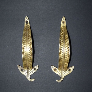 May include: Two gold-toned leaf-shaped door handles with intricate details. The handles have a pointed tip and a curved bottom.