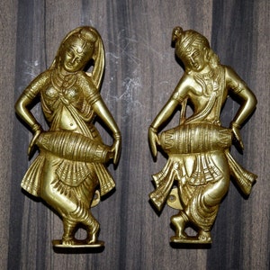 May include: Two golden brass sculptures depicting dancing figures with drums. The figures are in a dynamic pose, with detailed clothing and jewellery. The sculptures are mounted on a wooden surface.
