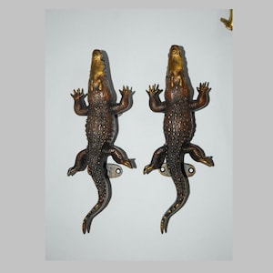 May include: Two decorative door handles shaped like crocodiles with gold accents. The handles are mounted on a white wall.