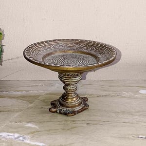 Puede incluir: Un plato de pedestal de latón antiguo con un diseño ornamentado y detallado. El plato tiene un cuenco redondo y poco profundo con un borde decorativo y un patrón central. La base del pedestal está intrincadamente tallada y el color general es latón antiguo cálido.