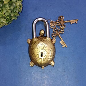 May include: A brass turtle-shaped padlock with a silver shackle and two decorative keys. The padlock has a detailed, textured design and is set against a blue background. The keys have an ornate, floral design.
