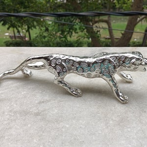 May include: A silver metal figurine of a leopard in a crouching position. The figurine is detailed with spots and has a realistic appearance.