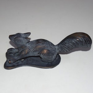 May include: A black and bronze metal squirrel door knocker. The squirrel is in a running pose with its tail raised. The knocker is mounted on a flat, oval-shaped base.